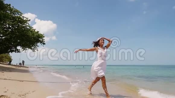 穿着白色连衣裙和花圈的迷人年轻快乐的女人光着脚沿着海岸奔跑享受着自己的生活视频的预览图