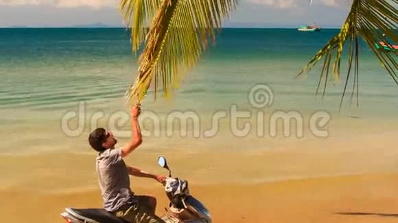 坐在摩托车上的男人微笑着抚摸着手掌下的棕榈枝视频的预览图