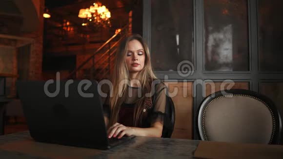 年轻女子坐在木桌上的咖啡店里喝咖啡用智能手机桌子上有笔记本电脑女孩视频的预览图