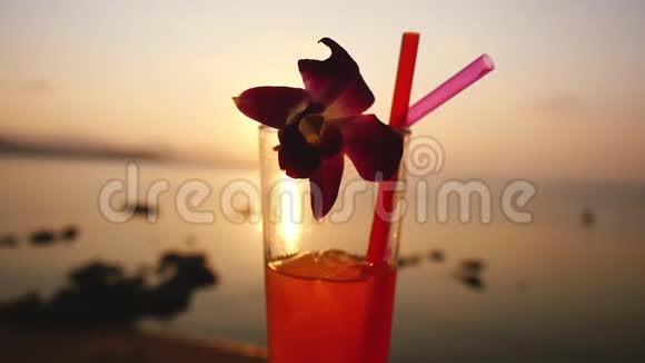 一杯热带鸡尾酒在令人惊叹的日落和美丽的海景背景下装饰了一朵花慢动作视频的预览图