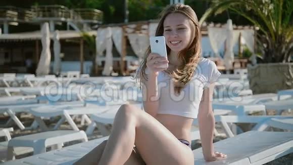 年轻女子在沙滩上躺在日光浴床上用手机视频的预览图