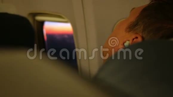 在飞机上休息的年轻人他睡在耳机里舷窗美丽的日出视频的预览图