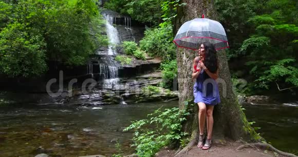 一个年轻的女人拿着雨伞站在瀑布边的森林里看着落水视频的预览图