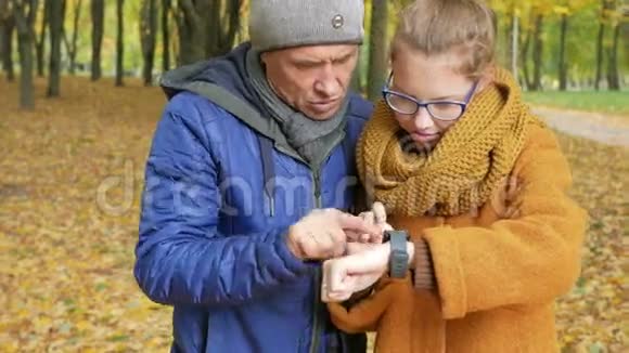 女儿少年教父亲在秋天公园里用智能手表工作爸爸仔细听重复一遍视频的预览图