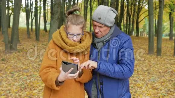 女儿少年教父亲在秋天公园里用平板电脑工作爸爸仔细听重复一遍视频的预览图