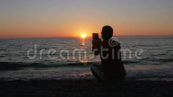 剪影男子与数码平板在日落海滩拍照太阳几乎落在海洋后面视频的预览图