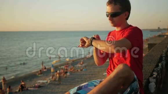 年轻的帅哥戴着太阳镜带着智能手表在海边放松他查看社交网络上的信息视频的预览图