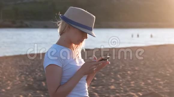 戴着帽子的漂亮金发女孩在日落时在海滩上的手机上键入一条信息视频的预览图