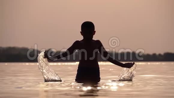 日落时快乐男孩的剪影用他的手创造了溅水慢动作视频的预览图