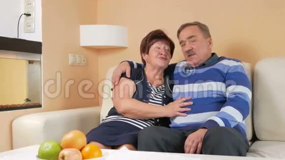 快乐的老人在一所漂亮的房子里的沙发上拥抱他心爱的妻子他们谈笑风生看着摄像机视频的预览图
