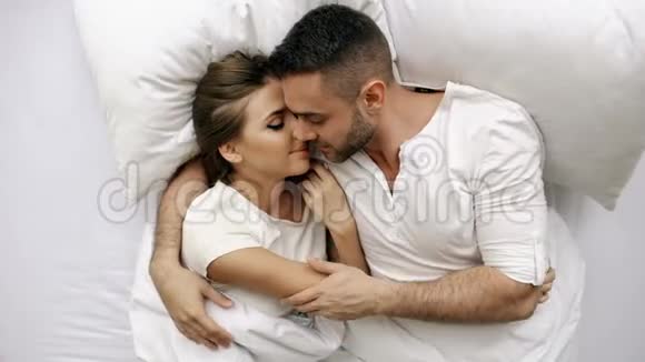 年轻美丽而有爱心的夫妇在早上醒来时亲吻并拥抱在床上迷人的男人的俯视图视频的预览图