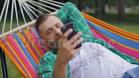一个长胡子的男人穿着绿色格子衬衫躺在吊床上用电话视频的预览图