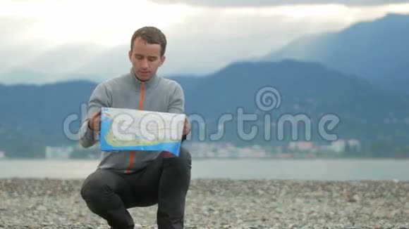 年轻英俊的男人坐在岩石海滩上它检查纸图上的方向在太平洋和视频的预览图