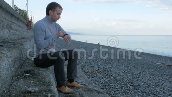 一个人坐在靠近海洋卵石海滩的台阶上它扫描智能手表上的信息并回应社交视频的预览图