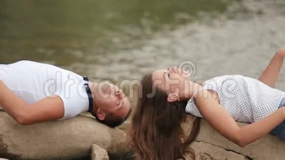 美丽的年轻夫妇躺在石岸上的大河边用手抚摸视频的预览图