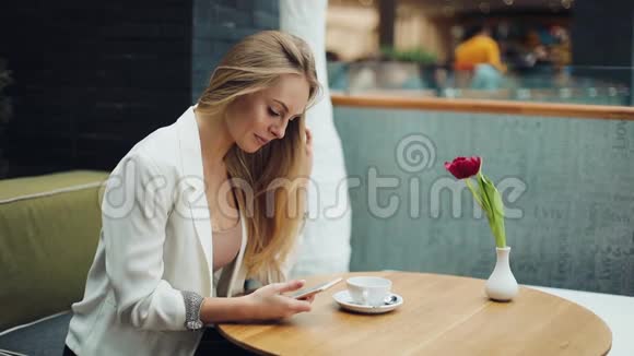 迷人的金发女人在她的智能手机上读到一些东西坐在咖啡馆的桌子旁视频的预览图