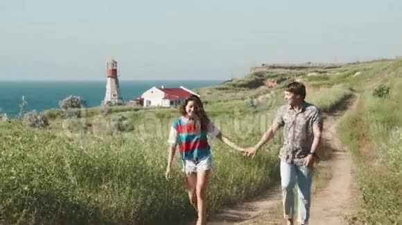 年轻的一对夫妇走在海滩悬崖上的灯塔附近的沙滩上谈笑风生慢慢视频的预览图