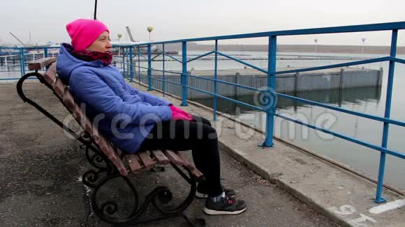 在寒冷的阴天女孩坐在码头上的长凳上微笑着视频的预览图