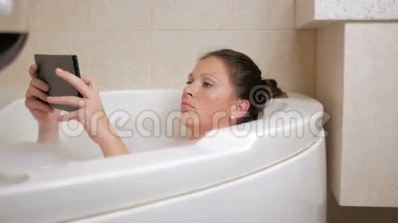 美丽的女孩洗澡与泡沫和平板电脑一个大白浴和一杯红酒盘子上的薄片视频的预览图