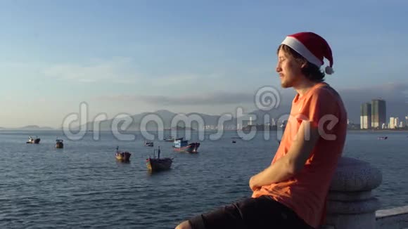 戴着圣诞帽的人坐在栏杆上看着大海中的钓鱼船升起了太阳视频的预览图