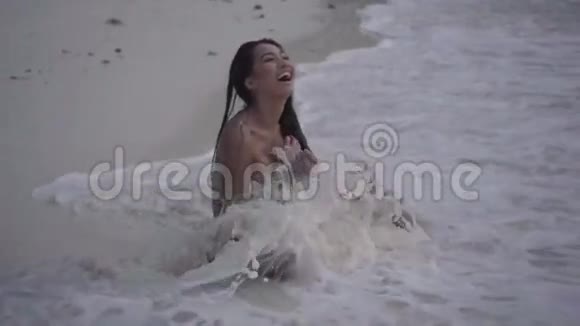 微笑的女孩坐在海滩上缓慢地玩波浪飞溅的视频视频的预览图