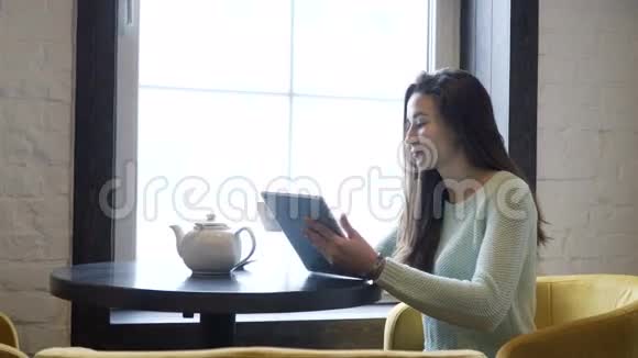 年轻的黑发女孩坐在靠近窗户的桌子上看着平板电脑喝茶视频的预览图