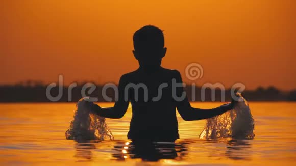 小男孩的剪影在夕阳下的河流创造飞溅的水与他的手慢动作视频的预览图