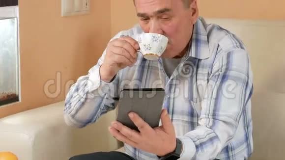 一位老人留着胡子在家里用平板电脑看新闻他坐在水族馆附近的沙发上喝咖啡视频的预览图