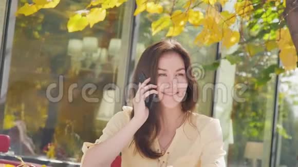 奇妙的秋景迷人的年轻女子开心地笑着打着电话在户外咖啡馆里拍照视频的预览图