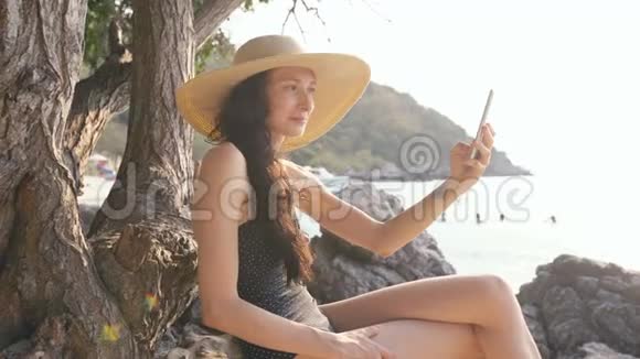在海滩度假期间穿着泳衣和帽子的年轻女子用智能手机拍摄自拍照片视频的预览图