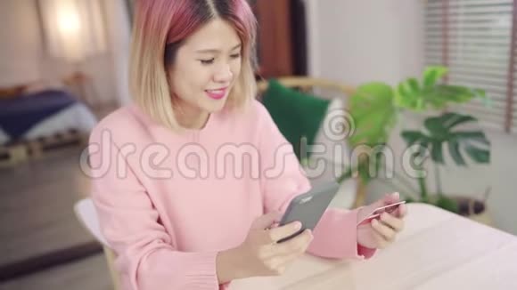 亚洲女性使用智能手机通过信用卡购买网上购物而穿毛衣坐在家里客厅的桌子上视频的预览图