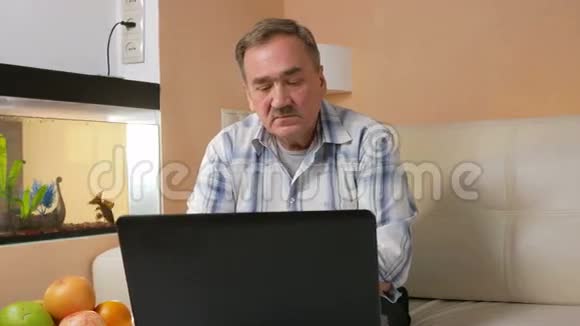 在家里的笔记本电脑上读新闻的老人胡子他坐在水族馆附近的沙发上盯着一台电脑视频的预览图