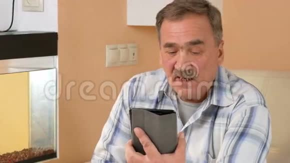 一位留胡子的老人在平板电脑上和亲戚交谈他坐在水族馆附近的沙发上视频的预览图