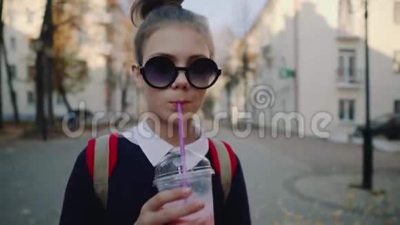 一个漂亮的红包时髦少年在建筑物之间的一条塑料杯步行街上喝奶昔可爱的女孩视频的预览图