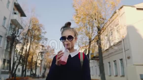 一个漂亮的红包时髦少年在建筑物之间的一条塑料杯步行街上喝奶昔可爱的女孩视频的预览图