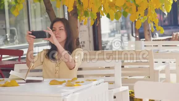 奇妙的秋景迷人的年轻女子开心地笑着打着电话在户外咖啡馆里拍照视频的预览图