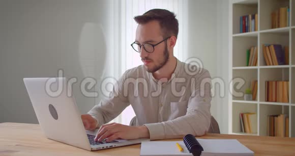 年轻英俊的白种人商人在笔记本电脑上打字在公寓的室内看着摄像机的特写照片视频的预览图