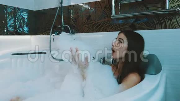 快乐的女孩在浴室里洗满泡沫的澡浴缸吹肥皂泡沫洗涤视频的预览图