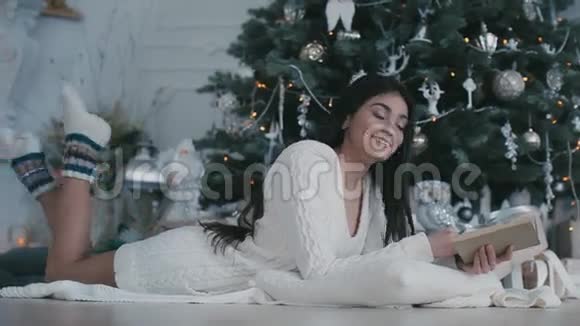 漂亮的女人躺在地板上的毛皮封面上靠近装饰的圣诞树看书视频的预览图