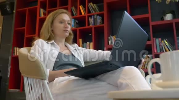 年轻女子微笑着坐在现代图书馆时使用笔记本电脑视频的预览图
