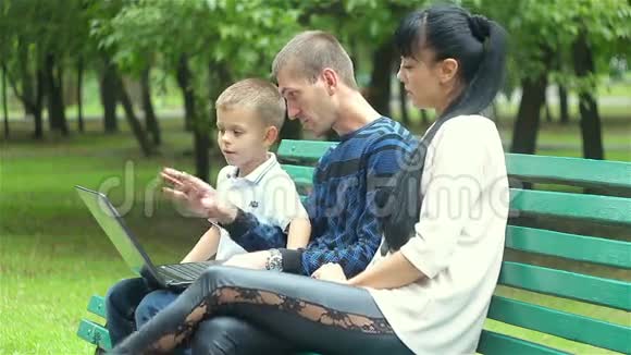 爸爸妈妈和儿子的家人坐在公园的长凳上看着一台笔记本电脑视频的预览图
