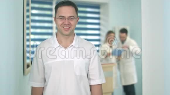 微笑男医生戴着眼镜看着摄像机而医务人员则在后台工作视频的预览图