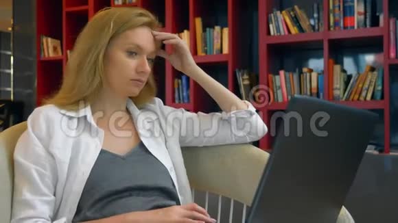 年轻女子微笑着坐在现代图书馆时使用笔记本电脑视频的预览图