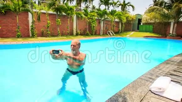 有胡子的老人站在游泳池里让赛尔夫在棕榈树视频的预览图