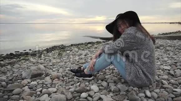 日落时分戴着黑帽子和眼镜的年轻美女坐在海边视频的预览图