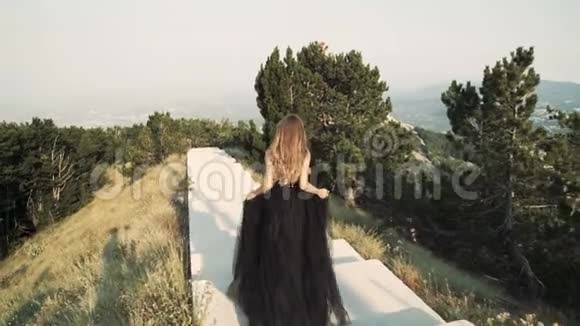 美丽的年轻女子模特穿着一件黑色蓬松的优雅长裙留着长发走在阳台上视频的预览图