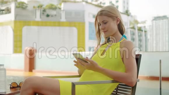 微笑快乐的女人穿着黄色的连衣裙坐在游泳池附近的椅子上她的智能手机视频的预览图
