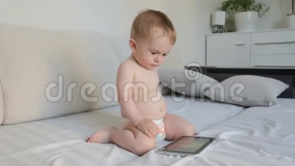 穿着尿布的可爱男婴坐在沙发上用数码平板电脑视频的预览图