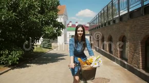 快乐的黑发女孩骑着自行车花和法国面包在篮子里微笑慢稳定的镜头视频的预览图
