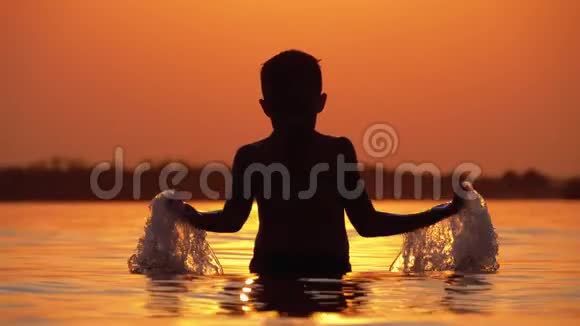 小男孩的剪影在夕阳下的河流创造飞溅的水与他的手慢动作视频的预览图